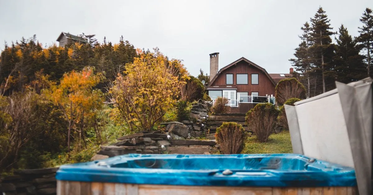 Blauer Whirlpool mit geöffneter Abdeckung in einem herbstlichen Garten vor einem Holzhaus